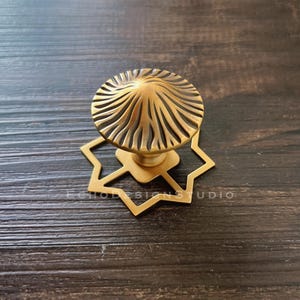May include: A brass-toned metal cabinet knob featuring a circular top with a radiating, textured sunburst pattern. The knob is mounted on a flat, star-shaped backplate with eight points. The hardware has a matte finish and a central stem.