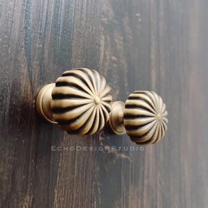 Może przedstawiać: Zbliżenie dwóch antycznych mosiężnych gałek do szafek z żłobionym, słonecznikowym wzorem. Gałki są zamontowane na ciemnej drewnianej powierzchni. Widoczne jest logo EchoDesignStudio.