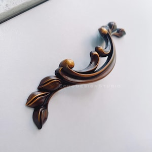 May include: Ornate bronze-colored cabinet pull with a floral design. The handle curves gracefully, featuring leaf-like details and a textured finish. The text "ECHODESIGNSTUDIO" is visible on the surface. This decorative hardware adds a touch of elegance to furniture.