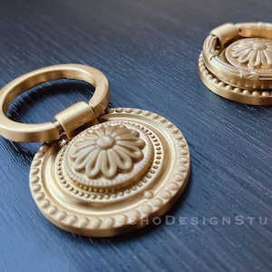 Könnte beinhalten: Zwei goldfarbene Metall-Schranktürgriffe mit verzierten Blumendesigns. Die Griffe haben einen ringförmigen Griff und eine runde Basis mit einem erhabenen Blumendesign.