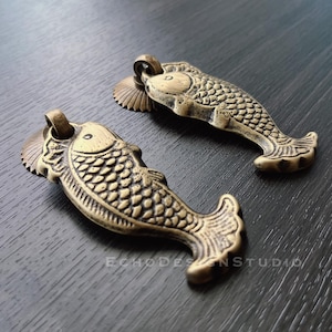 May include: Two gold-toned metal fish-shaped drawer pulls with intricate details. The pulls have a textured surface and a slightly raised design.
