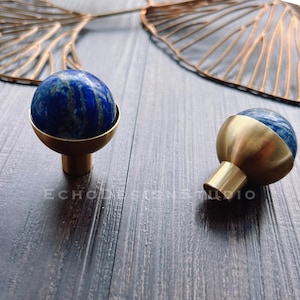 May include: Two round brass cabinet knobs with a blue lapis lazuli stone inlay. The knobs are on a dark wood surface.