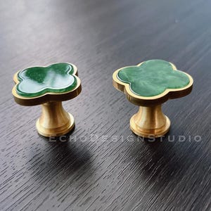 Könnte beinhalten: Zwei Messing-Schrankknöpfe mit einem grünen Kleeblatt-Design. Die Knöpfe haben eine glatte, polierte Oberfläche.