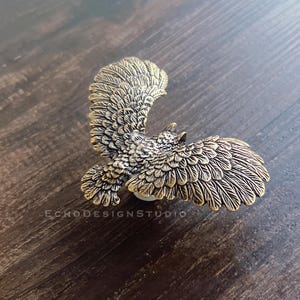 May include: A bronze-coloured cabinet knob shaped like an owl in flight. The knob has detailed feathering and is mounted on a white base. The text "ECHODESIGNSTUDIO" is visible on the surface.