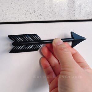 Könnte beinhalten: Schwarzer, pfeilförmiger Schrankgriff mit strukturierter Oberfläche. Der Pfeil zeigt nach rechts.