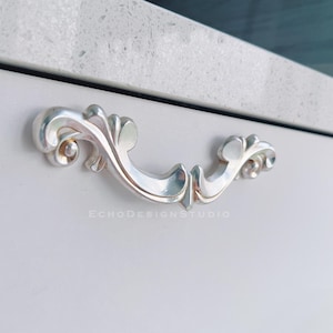 May include: A close-up shot of a silver drawer pull with an elaborate, curved design. The pull is fixed to a light grey surface. The text "ECHODESIGNSTUDIO" is visible below the pull.