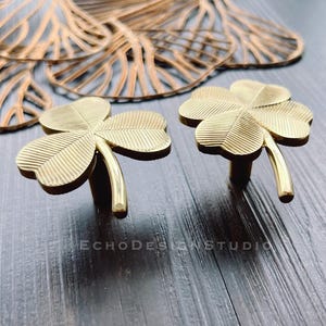 May include: Two gold-colored, four-leaf clover cabinet knobs. The knobs are made of metal and have a detailed leaf pattern. The background includes a decorative gold leaf design and a dark wood surface.