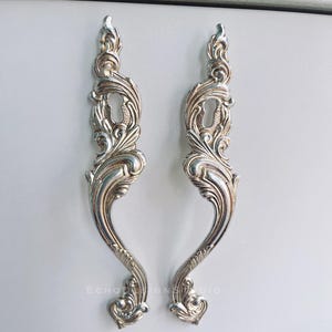 Könnte beinhalten: Zwei verzierte silberne Schrankgriffe mit Schlüssellochdetails. Die Griffe weisen ein geschwungenes, florales Design und eine geschwungene Form auf. Die Griffe sind wahrscheinlich aus Metall und für eine Vintage- oder Antik-Ästhetik konzipiert.