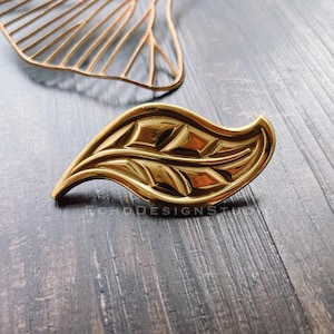 May include: A gold leaf-shaped decorative hardware with a detailed design. The hardware is made of metal and has a shiny finish.