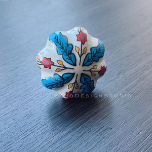 Puede incluir: Pomo de armario de cerámica blanca con un diseño floral azul y rojo. El pomo tiene un borde festoneado.