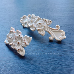 May include: Two white ceramic drawer pulls with gold accents. The pulls are shaped like flowers with intricate details.