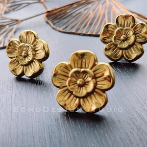 May include: Three gold metal flower-shaped drawer pulls. The pulls have a raised design and a slightly textured surface.