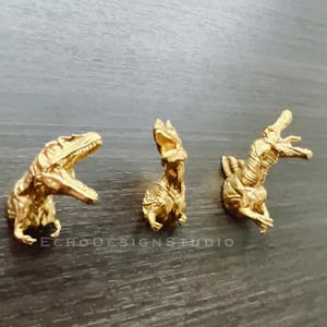 May include: Three gold-colored dinosaur figurines mounted on a wall. The dinosaurs are a velociraptor, a stegosaurus, and a tyrannosaurus rex.