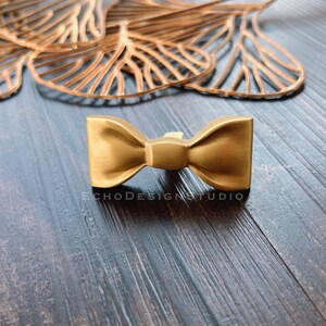 May include: Gold-toned bow tie shaped cabinet knob or drawer pull. The knob is made of metal and has a smooth, polished finish.