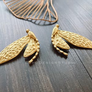 Könnte beinhalten: Ein Paar goldfarbene Ohrringe in Form von Libellen mit filigranen Details. Die Ohrringe sind aus Metall und haben eine strukturierte Oberfläche.