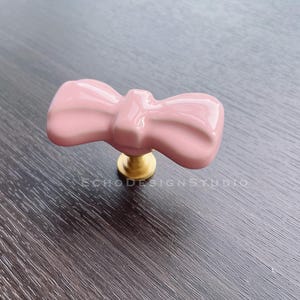 Op de afbeelding: Een roze keramische kastknop in de vorm van een strik met een gouden metalen basis.