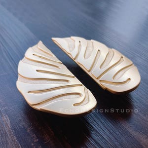 May include: Two gold-toned metal cabinet pulls shaped like a tropical leaf with a textured surface.