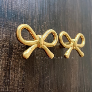 Könnte beinhalten: Zwei goldfarbene, schleifenförmige Schrankgriffe. Die Griffe bestehen aus glänzendem Metall und sind auf einer dunkelbraunen Holzoberfläche montiert. Der Text "ECHODESIGNSTUDIO" ist sichtbar.