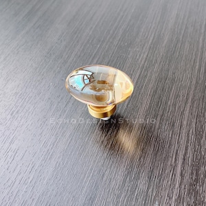 May include: A gold-toned cabinet knob with a clear glass oval top. The glass has a rippled texture and reflects the surrounding environment.