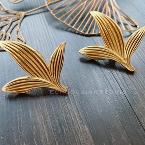 May include: A pair of gold leaf-shaped earrings with a textured design. The earrings are on a dark wood surface.