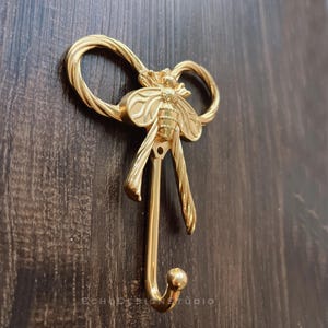 May include: A gold-coloured wall hook with a bee design. The hook features a detailed bee with outstretched wings, a bow-shaped top, and a single hook. The hook is mounted on a dark brown wooden surface. The text "ECHODESIGNSTUDIO" is visible.