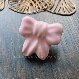 Puede incluir: Un pomo de cerámica rosa en forma de lazo. El pomo es un acento decorativo para muebles o armarios.