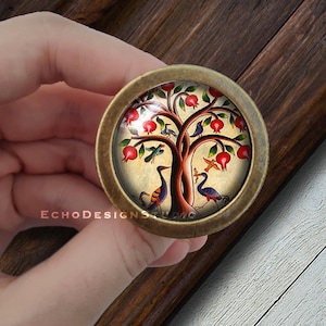 May include: A round, brass-colored knob with a glass insert featuring a colorful illustration of a tree with red pomegranates and birds. The tree is on a cream-colored background.