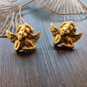 May include: Two gold-tone cherub angel earrings with wings and a thoughtful expression.