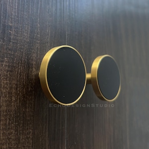 Puede incluir: Un par de tiradores de armario con un diseño moderno. Cada tirador presenta una cara negra y circular enmarcada en un marco de metal dorado. Los tiradores están montados sobre una superficie de madera oscura.