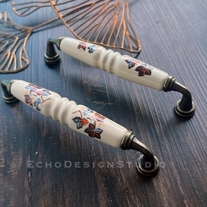 May include: Two ceramic cabinet knobs with a floral design. The knobs are white with brown and orange floral accents. The knobs have a brass finish.