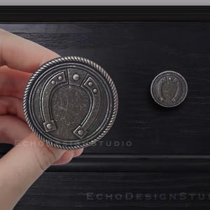 Puede incluir: Dos pomos de armario de metal plateados con un diseño de herradura. Los pomos tienen un borde elevado texturizado y un acabado ligeramente desgastado.