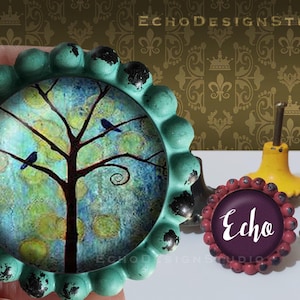 May include: Teal blue knob with a raised edge and a glass insert featuring a whimsical tree with birds and a blue, green, and yellow background. The knob is labeled "Echo".