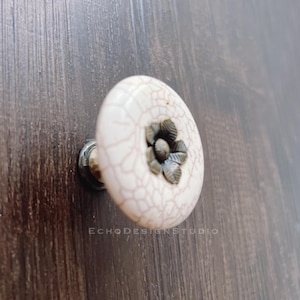 Peut inclure: Bouton de porte en céramique blanche avec une finition craquelée et un motif de fleur en métal au centre.