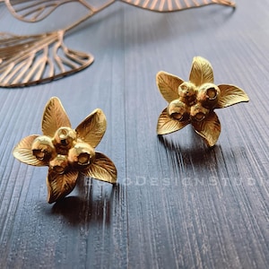 Peut inclure: Une paire de boucles d'oreilles à clous dorées en forme de fleurs. Chaque boucle d'oreille présente un motif floral à cinq pétales et un groupe d'éléments en forme de baies au centre. Les boucles d'oreilles sont posées sur une surface sombre imitation bois.