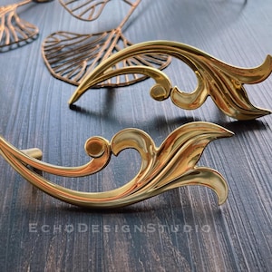 May include: Two gold-toned metal drawer pulls with ornate, swirling designs. The pulls are shaped like stylized leaves.