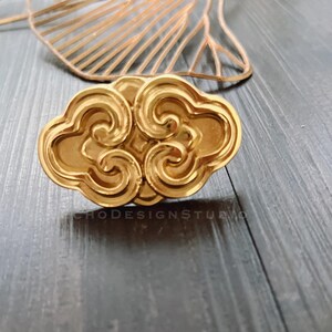 Może przedstawiać: Złoty, dekoracyjny przedmiot w kształcie chmury z wirującymi detalami. Obiekt umieszczony jest na szarej drewnianej powierzchni. Tekst "CHODESIGNSTUDIO" jest widoczny na dole obiektu.