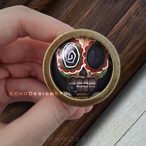 May include: A round, gold-colored metal pin with a colorful sugar skull design. The skull has black eyes, a red and white mouth, and a green and white floral design around the eyes.