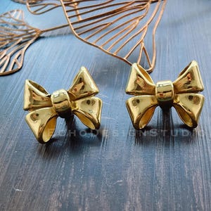 Peut inclure: Une paire de boucles d'oreilles en forme de nœud de couleur or. Les boucles d'oreilles sont en métal et ont une finition brillante.