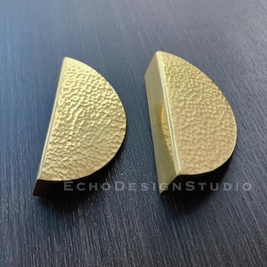 May include: Two half-moon shaped cabinet pulls in a hammered gold finish. The pulls are made of metal and have a textured surface. The pulls are displayed on a dark blue surface. The text "ECHO Design Studio" is visible.