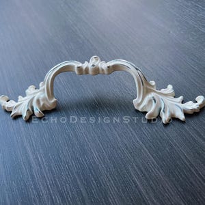 May include: White ornate cabinet pull with a decorative leaf design. The pull is made of metal and has a smooth, polished finish.