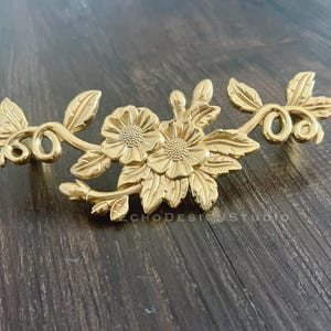 Op de afbeelding: Goudkleurige metalen ladegreep met een bloemmotief. De greep heeft twee gestileerde bloemen met ingewikkelde details en bladeren.