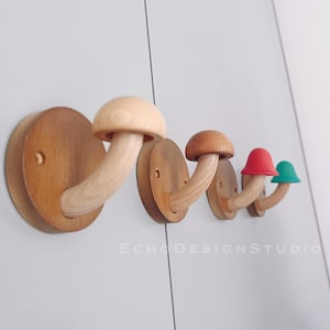Może przedstawiać: Cztery drewniane haczyki w kształcie grzyba zamontowane na ścianie. Haczyki są wykonane z naturalnego drewna i mają brązowe wykończenie. Haczyki są ułożone w rzędzie, z największym haczykiem po lewej stronie i najmniejszym haczykiem po prawej stronie. Haczyki są unikalnym i dekoracyjnym sposobem na dodanie miejsca do przechowywania w każdym pomieszczeniu.
