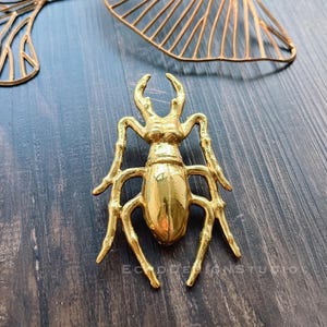 Puede incluir: Broche dorado con forma de escarabajo, con detalles en patas, antenas y segmentos corporales. El broche está muy pulido y refleja la luz. El fondo incluye una superficie de madera y un elemento decorativo dorado en forma de hoja.