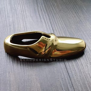 May include: A gold-colored shoe-shaped trinket or decorative object. The shoe is made of metal and has a shiny, polished finish.