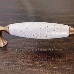 以下が含まれることがあります： 幾何学模様と金色の金属製のアクセントが付いた白いセラミック製のキャビネットノブ。ノブの長さは 12.7 cm です。