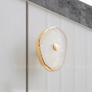 Puede incluir: Pomo de armario de ágata blanca con un borde de metal dorado y un pequeño pomo de metal dorado en el centro.