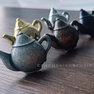 Pode incluir: Uma coleção de miniaturas de bules em vários acabamentos metálicos, incluindo dourado, prateado, bronze e preto. Os bules estão dispostos sobre uma superfície de madeira, mostrando seus detalhes intrincados e design clássico. O texto "ECHODESIGNSTUDIO" é visível.