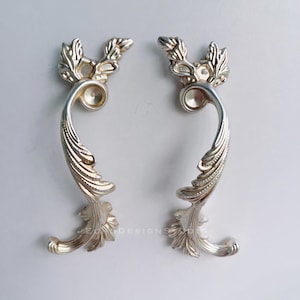 May include: Two silver-colored, ornate cabinet pulls. Each pull features a curved handle with a detailed leaf design and a decorative top. The pulls are designed with a vintage aesthetic, perfect for adding a touch of elegance to furniture.