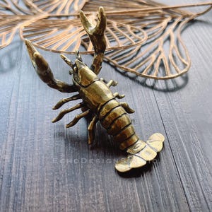 May include: A detailed brass crayfish figurine with raised claws and segmented body. The crustacean is positioned on a dark wood surface, with a decorative gold leaf in the background. The figurine is a detailed representation of a crawfish.