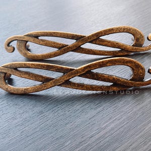 Może przedstawiać: Dwa uchwyty do szafek w kolorze brązu z motywem symbolu nieskończoności. Uchwyty mają teksturowane wykończenie i zakrzywiony, zwijany kształt na każdym końcu. Okucia do mebli i wystroju domu.